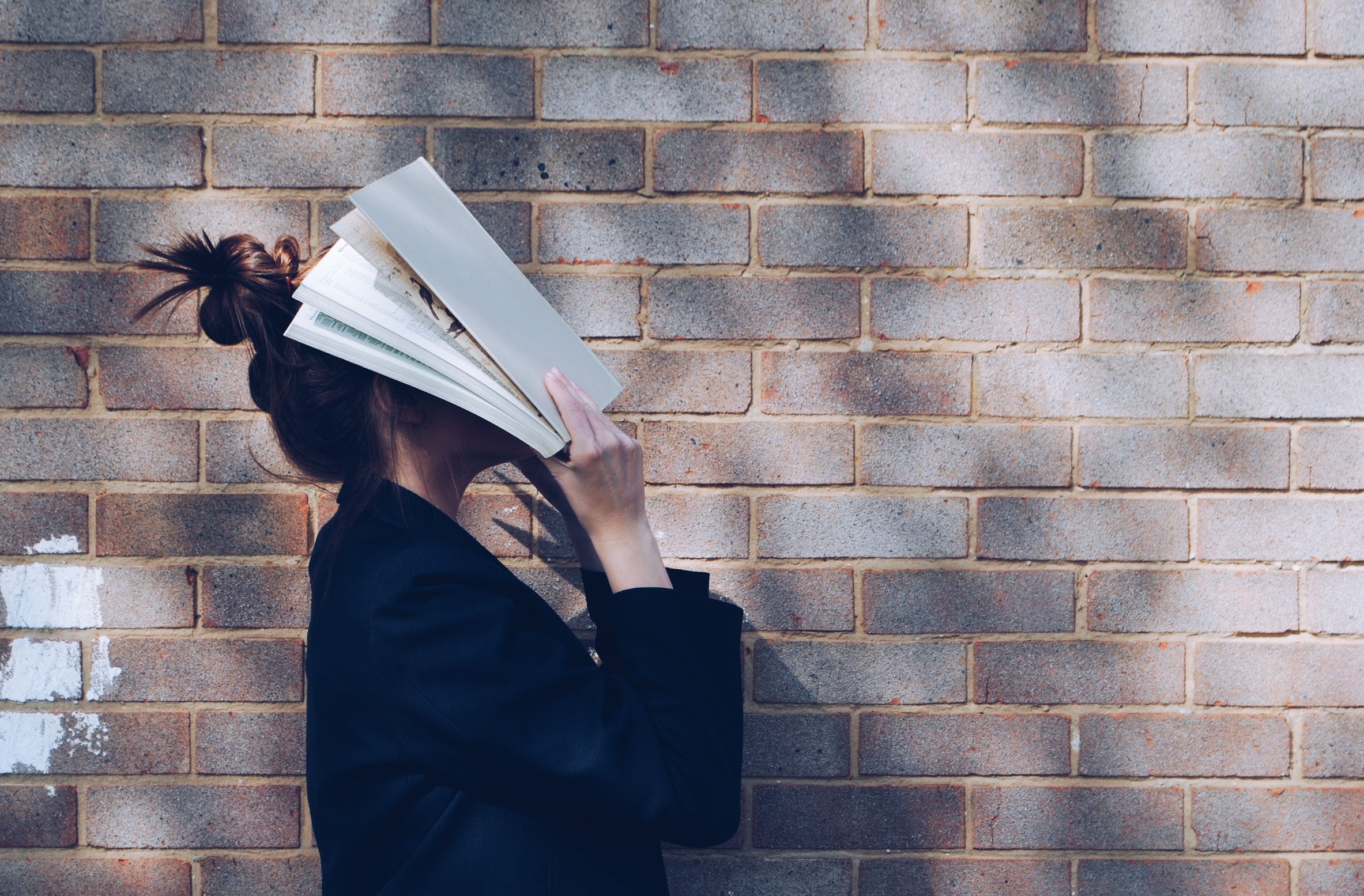 Woman covering her face with a book next to a brick wall